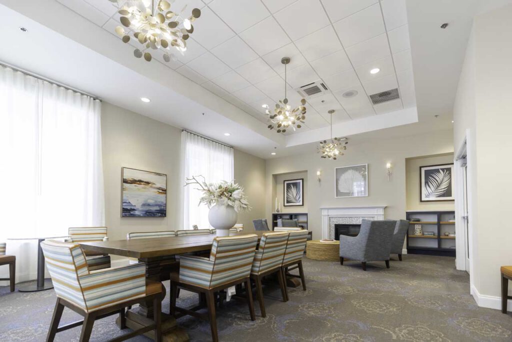 Bright, modern lounge with striped chairs around a wooden table, soft armchairs near a white fireplace, large windows with sheer curtains, decorative plants, and framed artwork on the walls. Contemporary chandeliers hang from the ceiling.