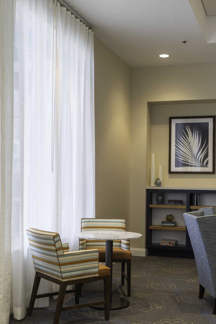 A small round table with two striped chairs sits near a tall window with sheer curtains. Behind the table is a bookshelf, a framed leaf print, and a light beige wall. The space looks modern and inviting.
