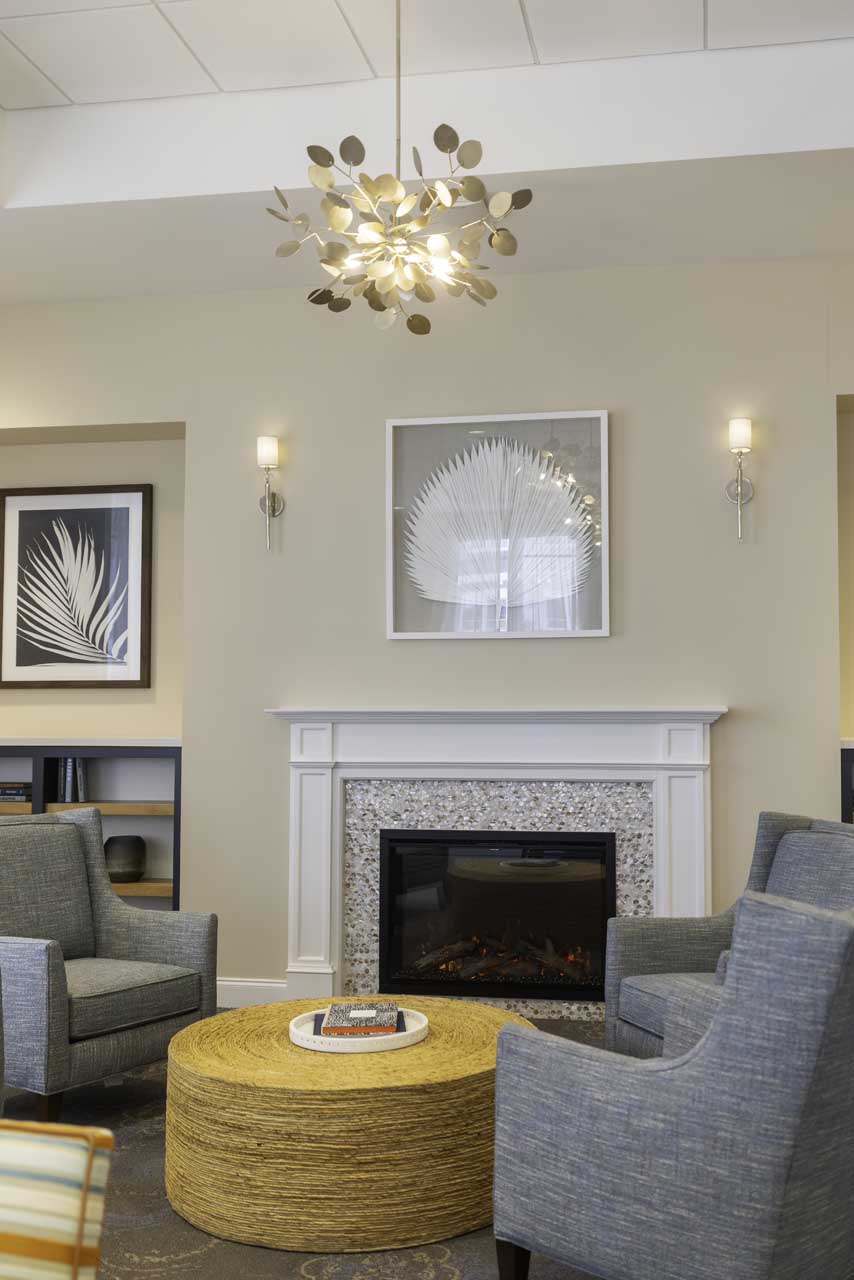 A modern living room with two gray armchairs, a round woven coffee table, and a fireplace. Above the mantel are two framed leaf artworks, and a contemporary light fixture hangs from the ceiling.