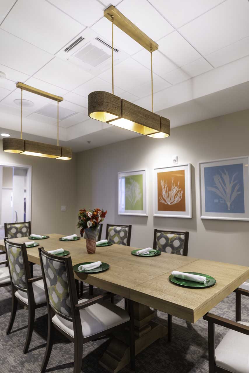 A modern dining room with a wooden table set for eight, featuring green plates and napkins, upholstered chairs, two pendant lights, wall art with botanical prints, and a vase of flowers as the centerpiece.
