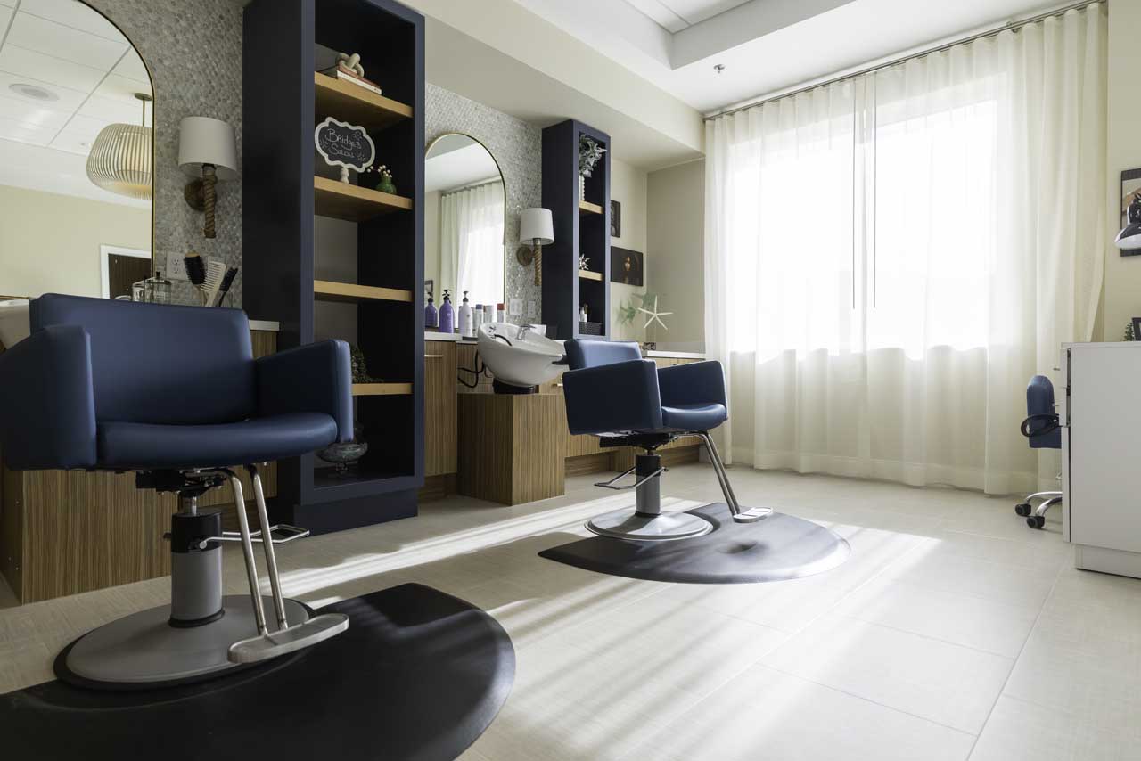 A bright, modern salon with two blue styling chairs, wooden shelves with products, a hair washing station, and large windows with sheer white curtains letting in natural light.