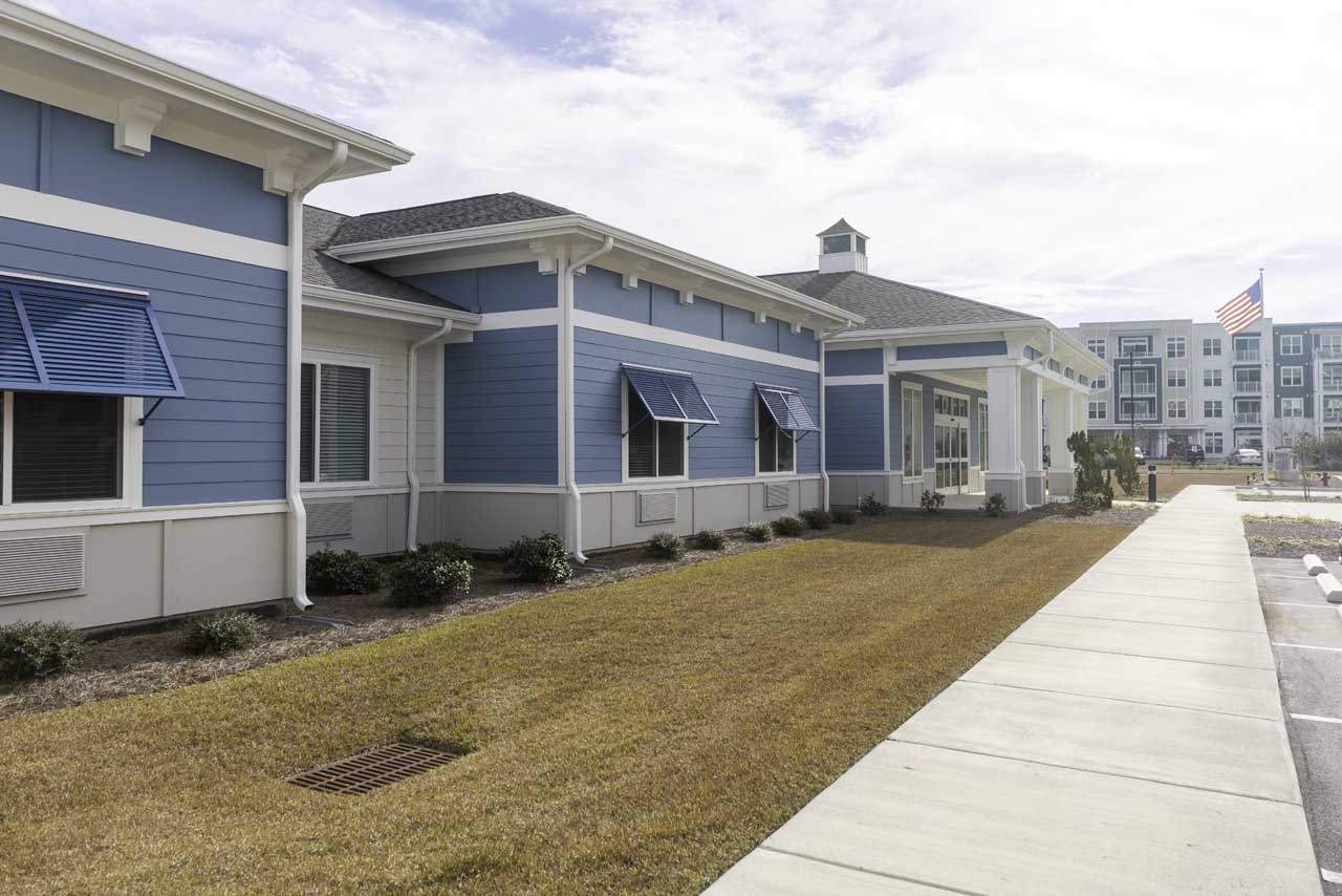 A modern blue and white building with blue awnings, landscaped bushes, and a neatly mowed lawn. An American flag is visible in the background near another building. The sky is partly cloudy.