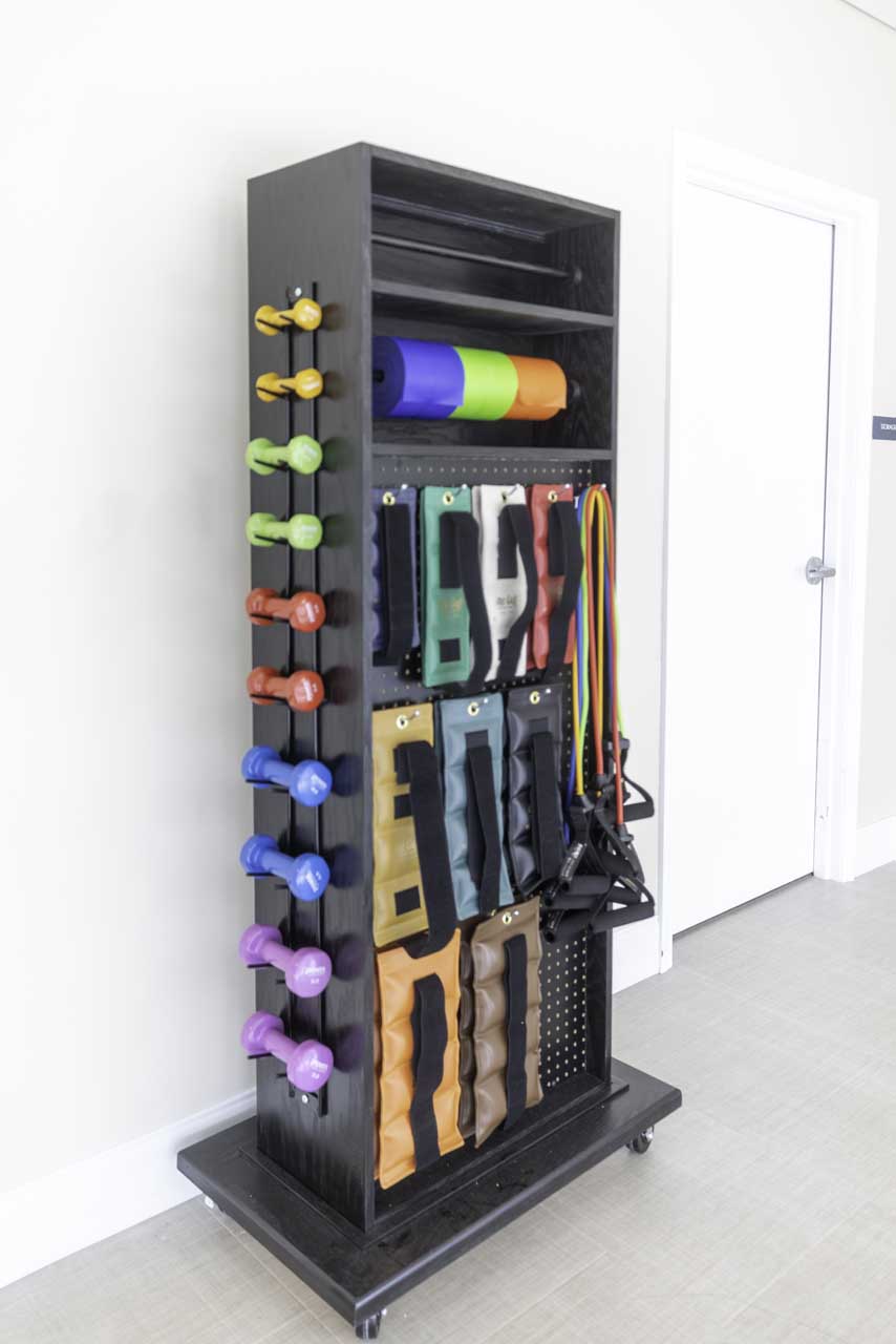A black storage rack holds colorful dumbbells, foam rollers, resistance bands, and exercise mats, organized neatly against a light-colored wall in a fitness or gym setting.