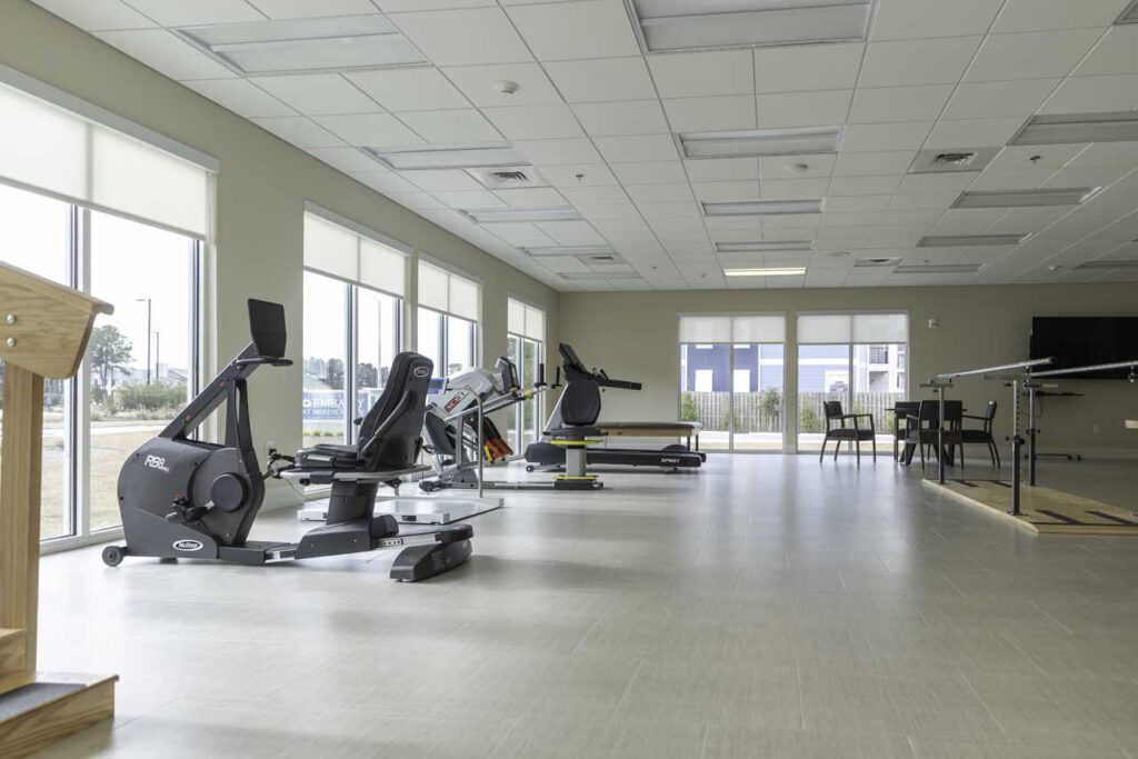 A bright, spacious gym room with large windows, exercise bikes, fitness equipment, a table with chairs, and a ping pong table. Light-colored floors and ceiling tiles create an open, airy atmosphere.