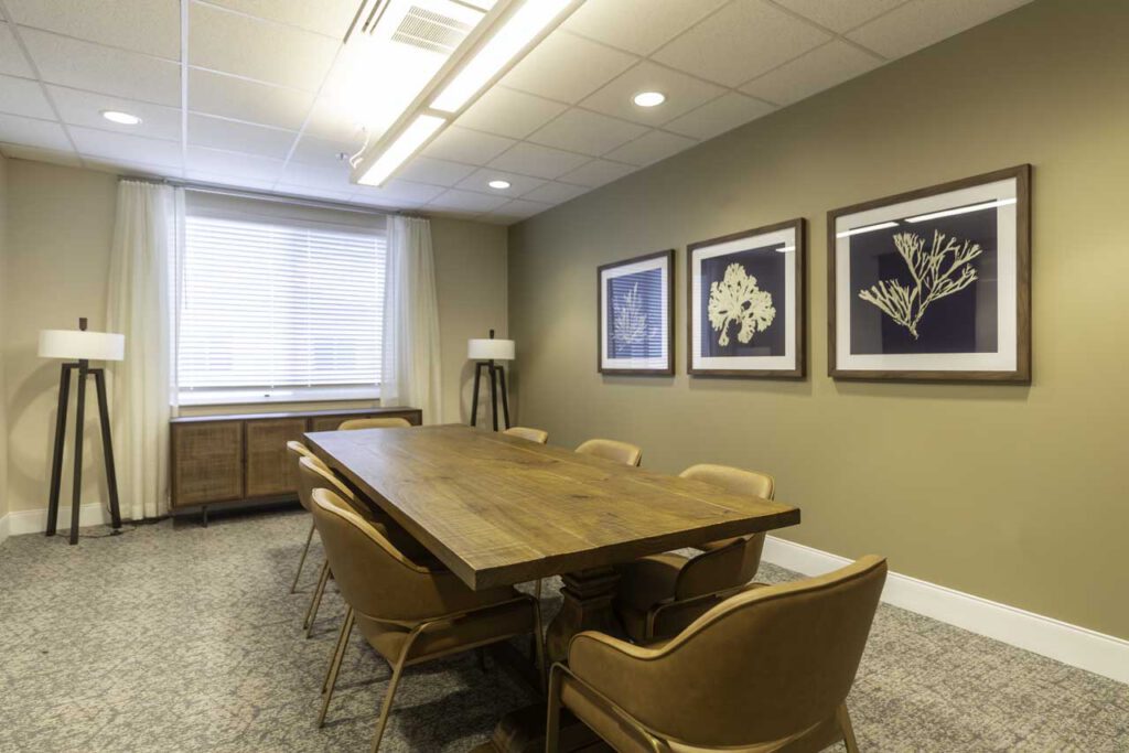 A small conference room with a wooden table, six brown chairs, beige walls, three botanical prints on the wall, two floor lamps, and a window with blinds letting in natural light.