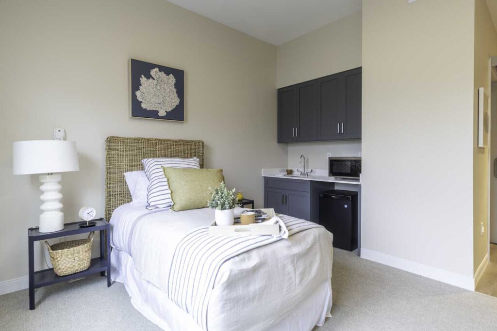 A cozy bedroom with a neatly made single bed, light bedding, a wicker headboard, a black nightstand with a white lamp, a mini kitchenette with gray cabinets, a sink, microwave, and mini fridge.