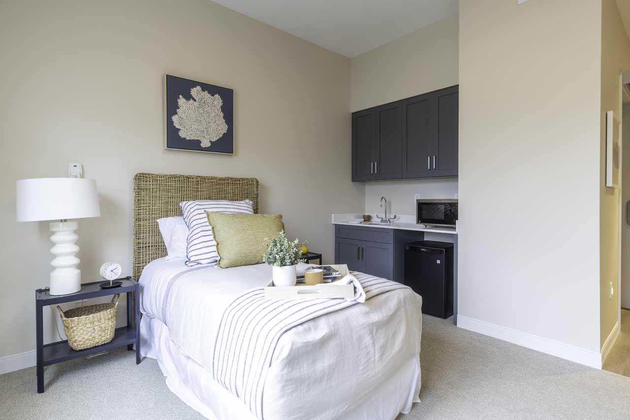 A cozy bedroom with a neatly made single bed, light bedding, a wicker headboard, a black nightstand with a white lamp, a mini kitchenette with gray cabinets, a sink, microwave, and mini fridge.