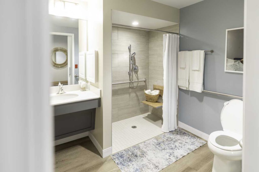 A bright, modern bathroom with an accessible walk-in shower, a curtain, grab bars, a toilet, a vanity with a mirror, white towels on racks, and a basket in the shower area. The floor has a patterned rug.