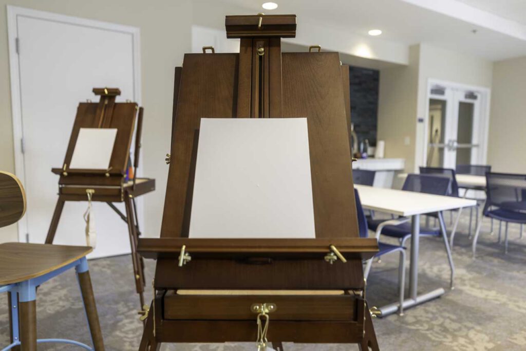 A close-up of a wooden easel holding a blank canvas in a well-lit room, with another easel and empty tables and chairs visible in the background.