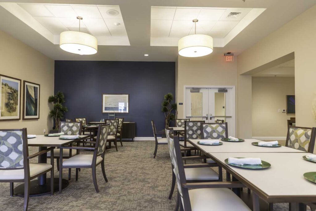 A modern dining room with neatly arranged tables and chairs, each table set with plates and napkins. The room has framed artwork, light fixtures, plants, and neutral-colored walls with a dark blue accent wall.