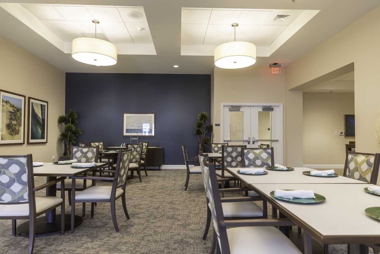 A modern dining room with neatly arranged tables and chairs, each table set with plates and napkins. The room has framed artwork, light fixtures, plants, and neutral-colored walls with a dark blue accent wall.