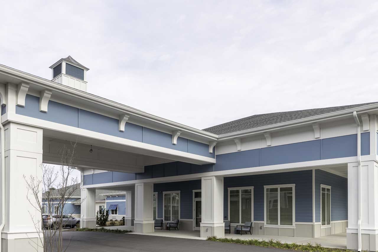 A modern building with light blue siding, white trim, and a covered driveway. Several windows and benches are visible along the exterior, and landscaped bushes line the front. The sky is overcast.