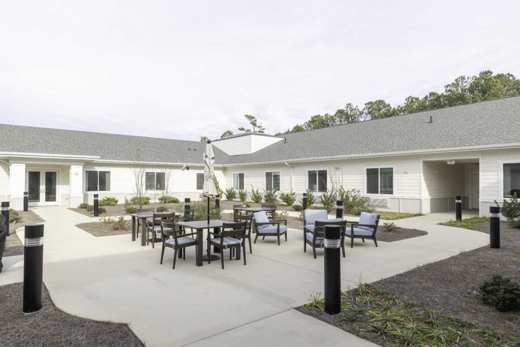 A courtyard with patio tables, chairs, and benches surrounded by a one-story building. The area has landscaping with shrubs and small trees, and the sky is clear.