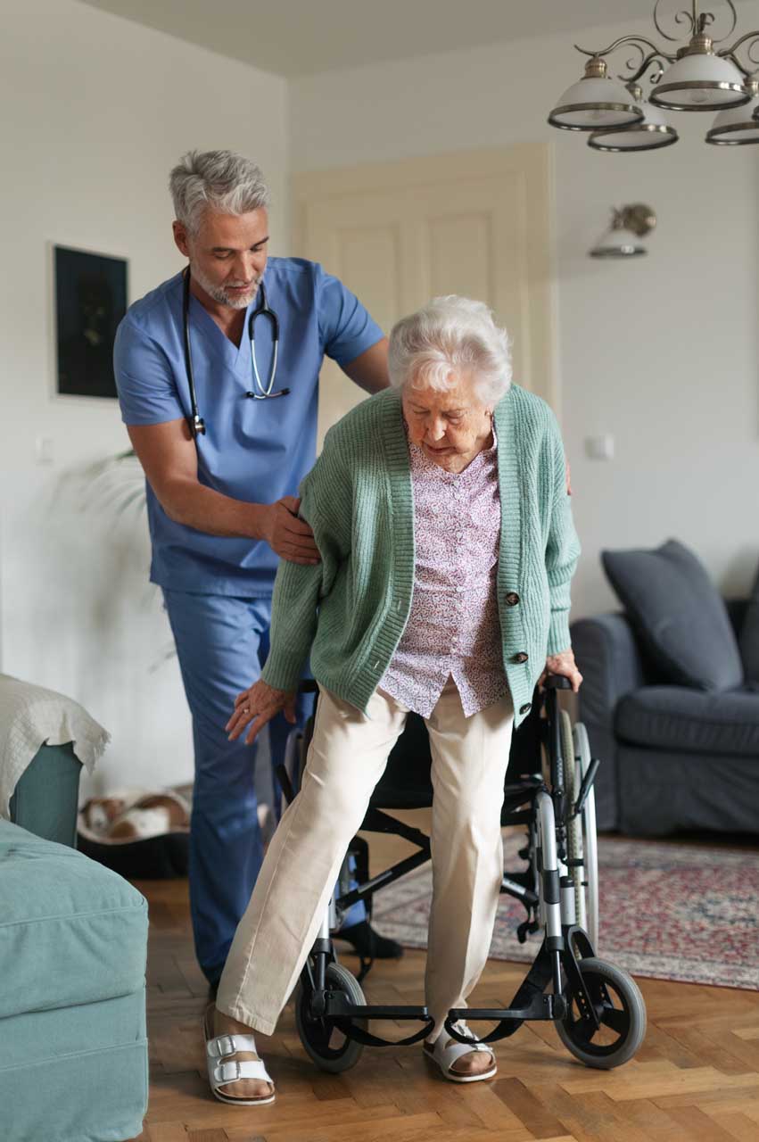 A healthcare professional in blue scrubs assists an elderly woman standing up from a wheelchair in a living room, offering support as she walks. The setting appears calm and supportive.